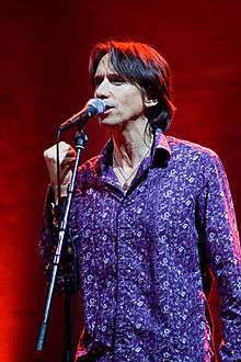 A Caucasian male with short medium brown hair. The male is wearing a purple buttoned down shirt with white designs spread across the shirt. He is standing in front of a microphone on a stand, while speaking into the microphone and clutching his right hand.