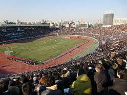 A packed oval shaped stadium with an athletics track. An urban skyline is seen in the horizon and the sky is blue with a few clouds.