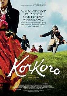 DVD cover for the movie, with a lady in Roma attire in the foreground, her face partially outside the frame, we can see her lips, chin and nose. In the background can be seen three men: one with grey hair, one with long black hair and the last with a hat on. The man with grey hair is shown about to jump. There is also a woman in the background, with not much of her in focus, except that she is wearing purple ethnic clothing.