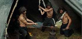 Three men are shown topless with black coloured pants on, beating hot iron. The man on the left holds the red hot iron in position while the one in the centre is shown about to strike, with the one on the right watches. The man in the centre is wearing a hat, while the other two sport unkempt hair. The man watching also has a towel on his shoulder. A girl is also in the picture, but hidden partially by the man on the left. The men have a slim muscular body with slim waists and are sweating profusely. We can see the well formed biceps of the man on the left who is striking the hot iron.
