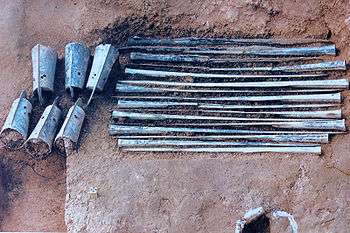 Pit with about ten long blue-green rusty metal sticks arranged parallel to each other and six bells.