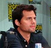 Color photograph of a white man with brown hair sitting in front of a ComicCon poster.