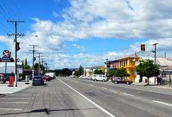 A small-town straight street, flanked by shops, parked cars and the occasional small tree. A Caltex petrol station is on the left, a two-storey pub painted bright blue and yellow is on the right.