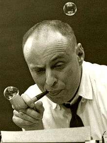 A black-and-white photo of a bald middle-aged man, blowing bubbles from a bubble pipe