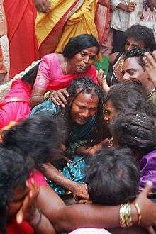 A group of seated transvestite men and/or eunuchs dressed in saris mourn.