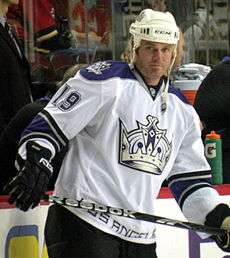 Hockey player in white uniform with a large crown in the middle. He holds a stick horizontally