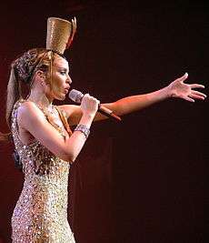 A woman is in a blue, sparkly showgirl outfit with a headpiece. She has a microphone and is singing.