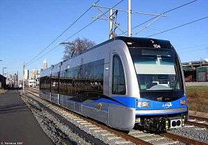 A Blue and gray train with black glass stands idle with overhead wires and adjoining building visible.