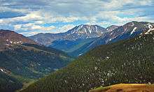 A view of mountainous terrain with a forested valley at the bottom rising to bare slopes with snowfields higher up. In the center one peak stands higher than the others.