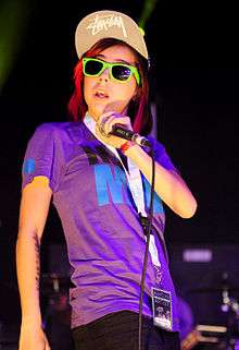 A woman in a purple t-shirt and black skinny jeans with sun glasses and a baseball cap with a "Bobbed" haircuit singing into a microphone