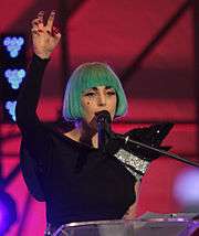 A woman wearing a blue bob wig, stands talking behind a podium. She raises her right hand in a claw like shape.