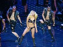 A blond woman wearing a black bustier embellished with shiny crystals performs next to two male back up dancers both wearing black leather jackets embellished with crystals and ripped jean shorts