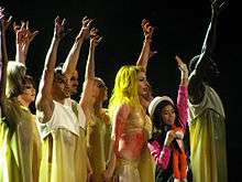 A grooup of dancers on stage wearing light yellow colored dress, and with their right hand stretched upwards in a claw shape. In their middle is a little girl wearing a pink dress and hat, and carrying a stuffed monkey.
