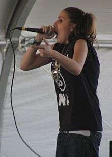 A Caucasian woman in her mid-twenties raps with a microphone to her mouth, wearing long brown pigtails, a black blazer, a yellow t-shirt, and a golden necklace.