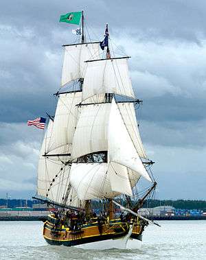 A wooden sailing vessel with numerous rope connecting from the deck to several large white sails above. An American flag drapes down from a pole at the back of the ship and a Washington state flag is on the tallest mast. The ship is in dark blue water, and in the background are a shoreline, treeline and cloudy sky.