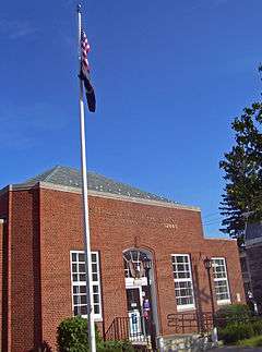 Three-quarter angle view of post office, a small brick building with hipped roof and a flagpole in front