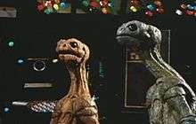 Two reptile-like alien creatures, one blue and one orange, look in an upward direction, with machinery and computer screens in the background.