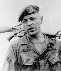 Head and shoulders of a white man wearing a t-shirt and a military jacket, unbuttoned at the collar, with the word "Rabel" on his right breast and "U.S. Army" on his left. He has on a beret and is holding a rifle over his shoulder by its barrel.