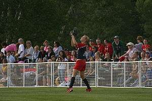 A light-skinned woman, wearing a red and blue shirt and red shorts, has her arms in the air to catch a ball in a grassy field as spectators look on.