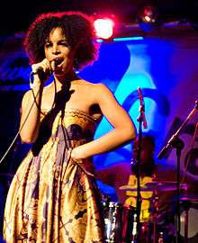 A woman sings into a microphone against a blue backdrop and red light. A man performs drums in the background.