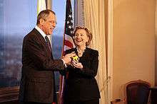 Hillary Clinton standing with Russian Foreign Minister Sergey Lavrov. Both of them are holding a "reset button". They are in a room with a window to the left and an American flag behind them