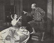 Woman kneeling in front of a standing man; the two are conversing amiably, and each is gesturing with one hand as if ringing a small bell, although, according to the song lyrics, they are actually demonstrating the flying motion of a honey bee