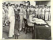 A large crowd of men in uniforms. Leclerc sits at a small desk signing a document. MacArthur stands behind him.