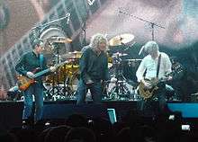 Led Zeppelin onstage: John Paul Jones playing bass guitar, Robert Plant holding a microphone, and Jimmy Page playing guitar. The trio stand in front of Jason Bonham's drum kit.