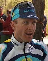 A profile shot of a man in his mid-thirties wearing a blue and white cycling jersey with yellow trim, and a matching cap, with a pair of sunglasses on top of the cap