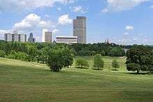 A rolling, grassy landscape with some trees beyond which some tall buildings can be seen under a blue sky with clouds. The tallest are light-colored modernist structures in the center of the image; smaller, older and darker buildings, including two church spires, rise above the treetops closer to the edges.