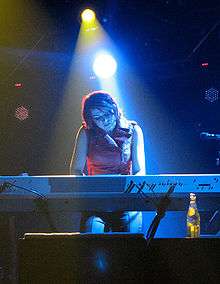 Lisa Harriton—a Caucasian woman with shoulder-length brown hair—plays keyboards onstage