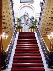 A red-carpeted staircase seen centrally from below with banisters on each side. At the top is a statue of a standing man wearing a toga and holding a scroll, and on each side are chandeliers on stands
