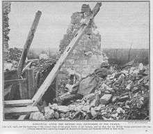 Monochrome image on newsprint type paper. Destroyed house with one remaining wall and visible roof timbers. Image of soldier dressed in British helmet and great-coat and rifle lying prone, peering over rubble towards the top right of picture.