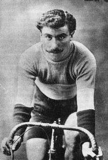 A black-and-white photograph of a man with three-colored sweater and shorts with a mustache sitting on a bicycle.