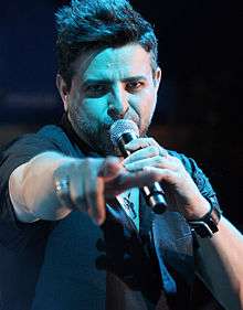 A man holding a microphone to his mouth with his right hand and pointing at the frotn with his left hand.