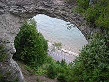An arch-shaped rock. The opening in the rock is sizable, and part of a road, trees, and a lake can be seen through the rock.