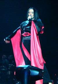 Madonna performing during the Drowned World Tour. She has a short brunette hair wig and wearing a black-and-red kimono. She is singing, while holding a microphone with her left hand