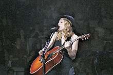 Image of a blond woman. She is dressed all in black but showing her arms, and wearing a hat of the same color. She is playing an orange-red acoustic guitar while a microphone is to her mouth. She is looking to her right and smiling