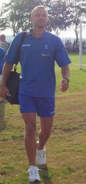 A muscular young white man wearing a blue sports shirt and shorts and carrying a kitbag walks across sunlit grass. In the background are trees and the edge of a football goal.