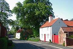 A pair of whitewashed cottages, with pantiled roofs, widely spaced along a road.  the nearer one has a red gate and a small pantiled outhouse.  A huge oak, in full leaf, stands behind the further one.