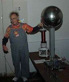  A man wearing coveralls with his left hand near the metal sphere of a Van de Graaff generator. His hair is standing on end due to electrostatic repulsion.