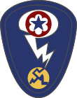 Oval shaped shoulder patch with a deep blue background. At the top is a red circle and blue star, the patch of the Army Service Forces. It is surrounded by a white oval, representing a mushroom cloud. Below it is a white lightning bolt cracking a yellow circle, representing an atom.