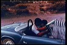 A frame of a man and woman kissing in a convertible sports car; the clapperboard is visible in the edge of the frame