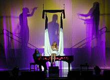 A blonde woman sits atop a piano and sings. A long, white cloth hangs around her while a silhouette behind the woman shows two male figures as if holding the white cloth.