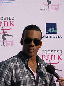 A black man, wearing sunglasses and a chequered shirt, speaks into a microphone whilst standing in front of a backdrop covered in sponsors' logos.
