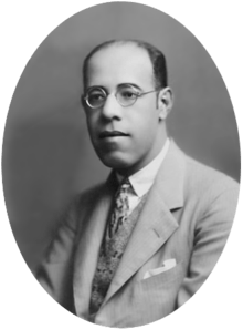 Photograph showing the head and shoulders of a man with glasses wearing a suit
