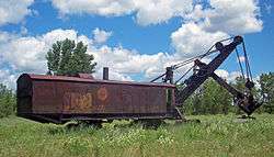 A brown rusted metal structure resembling a railroad boxcar with a rounded roof sitting in an overgrown field. On its right is a two-part shovel mechanism with gears, chains and a dipper at the end.