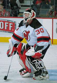 A man in full hockey goaltender equipment: mask combining helmet and cage, large gloves on the hands, leg pads atop his pants, and a wide stick on his right hand.