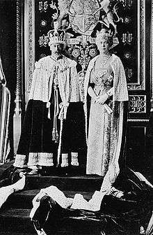 Late-middle-aged George and Mary in crowns and ermine capes stand on a dais