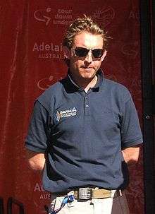 A blond man in his late thirties wearing a dark blue polo shirt and sunglasses, with his hands behind his back such that no part of his arms below either elbow is visible.